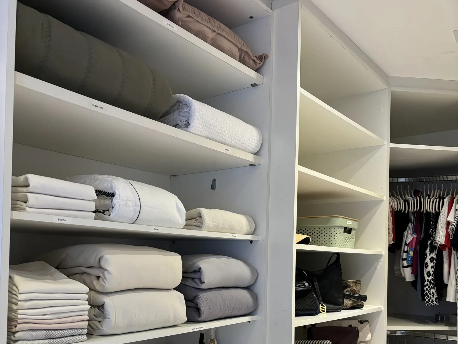 Rouparia branca com roupas de cama dobradas por categoria e etiquetas na borda das prateleiras